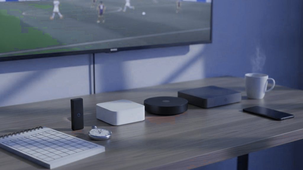 Five streaming devices, a notebook scoring grid and a stopwatch in front of a TV showing a football match